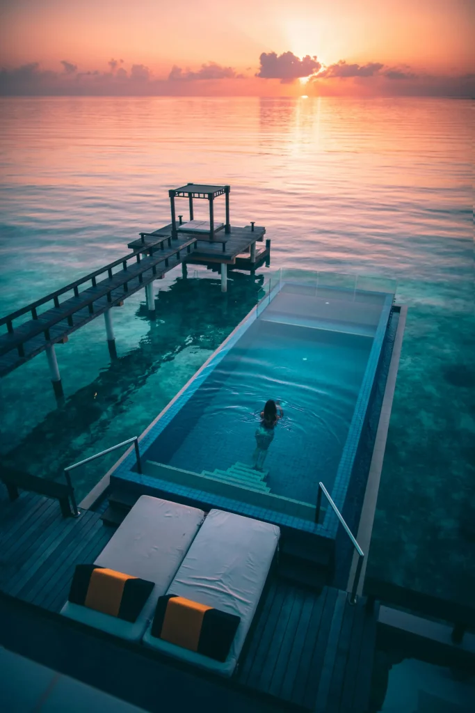 A person relaxes in an oceanfront pool beside a wooden jetty as warm orange light reflects across the turquoise sea.
