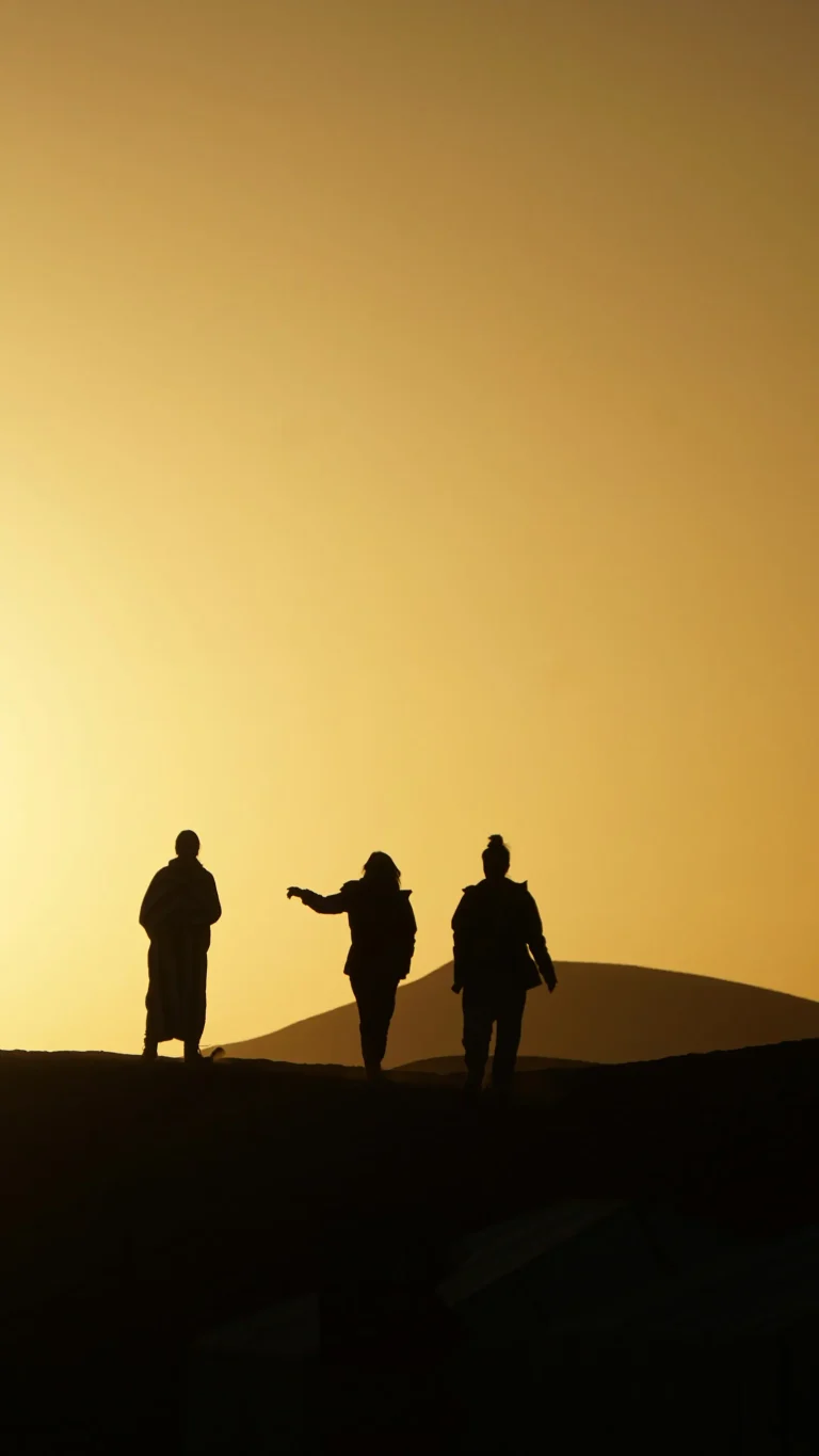 Three people silhouetted on a dune against an amber sky