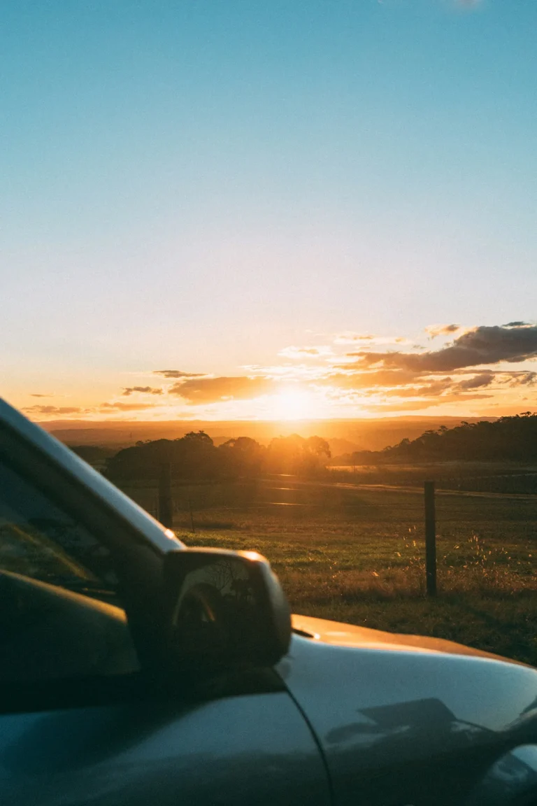 Car wing mirror in the foreground with the sun setting over open fields